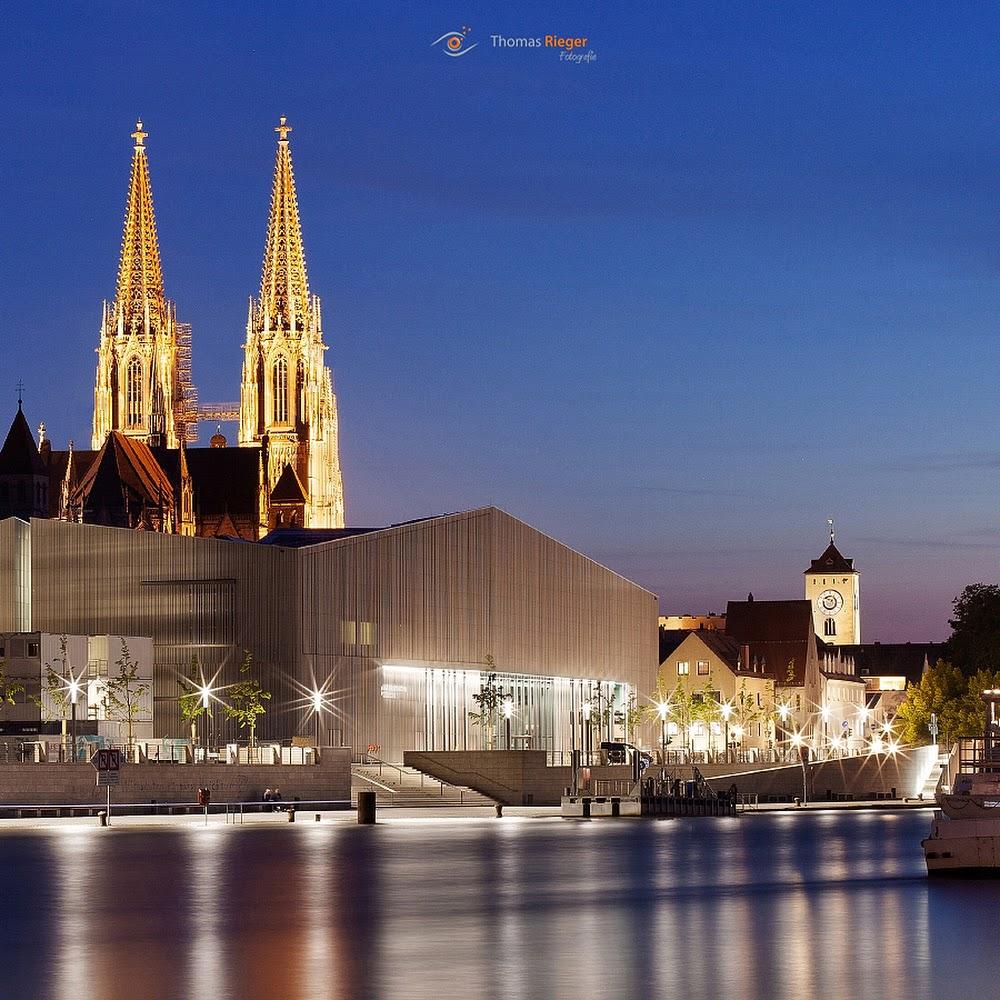 A church with two towers, a modern building, and water. The landmark is the Regensburg Cathedral.